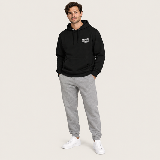 A man modeling a black pullover hoodie with a small white embroidered logo on the left chest, paired with heather gray jogger sweatpants and white sneakers, against a solid light beige background.