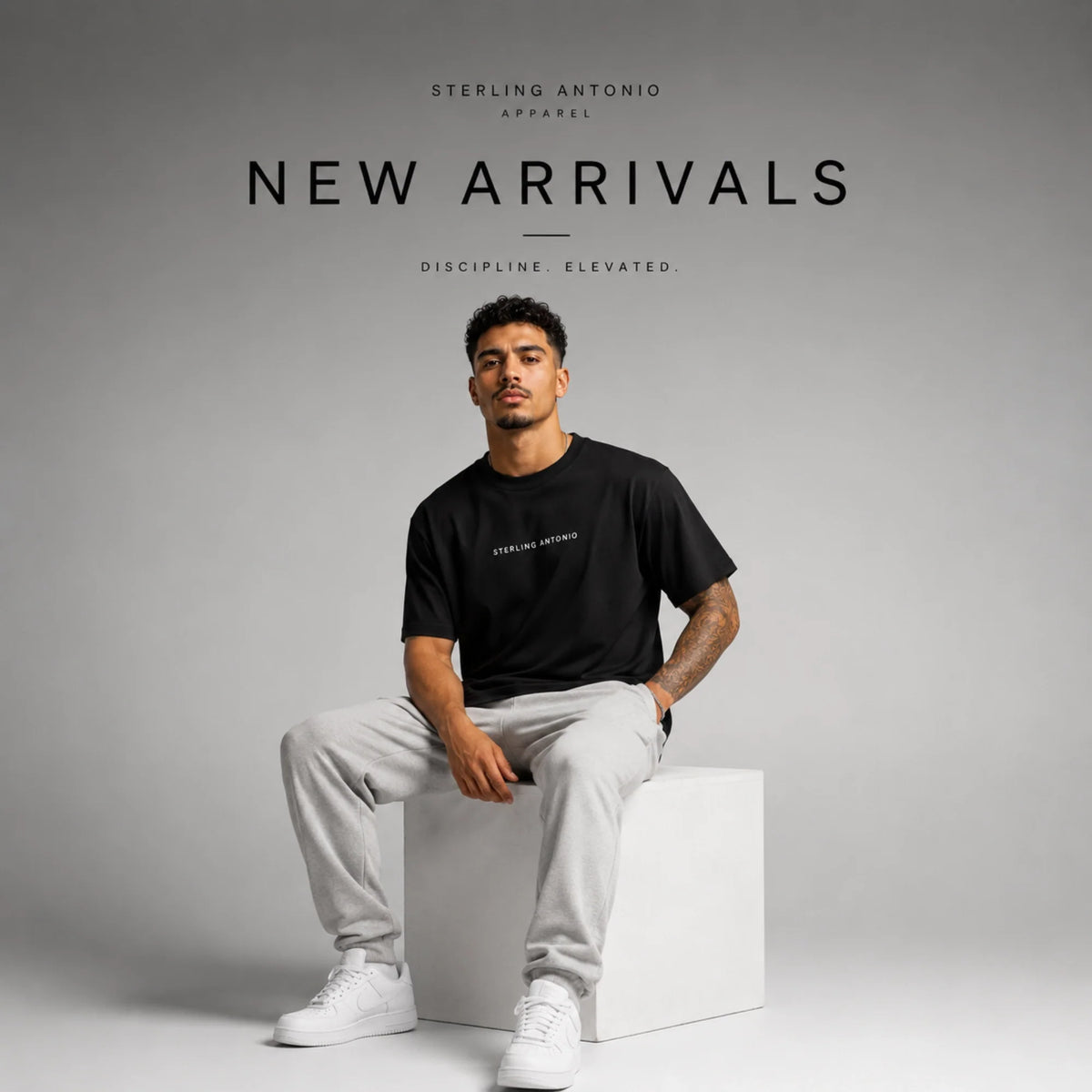 Male model wearing a black Sterling Antonio T-shirt with gray sweatpants seated in a modern studio, representing the New Arrivals collection of premium luxury streetwear.