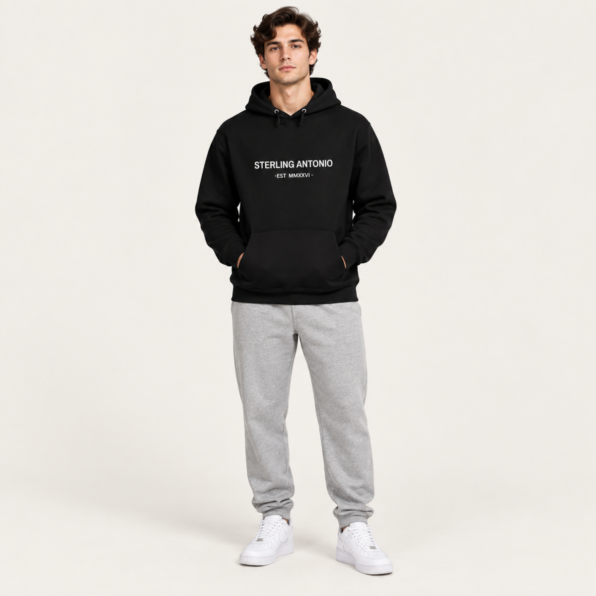 A man modeling a black pullover hoodie with 'STERLING ANTONIO EST. 2024' printed in white on the chest, paired with light gray sweatpants and white sneakers, against a plain off-white studio background.