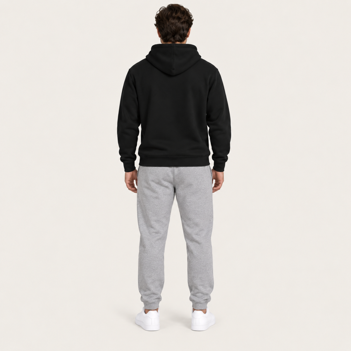 A man viewed from behind wearing a black pullover hoodie and light gray sweatpants, standing against a plain off-white studio background.