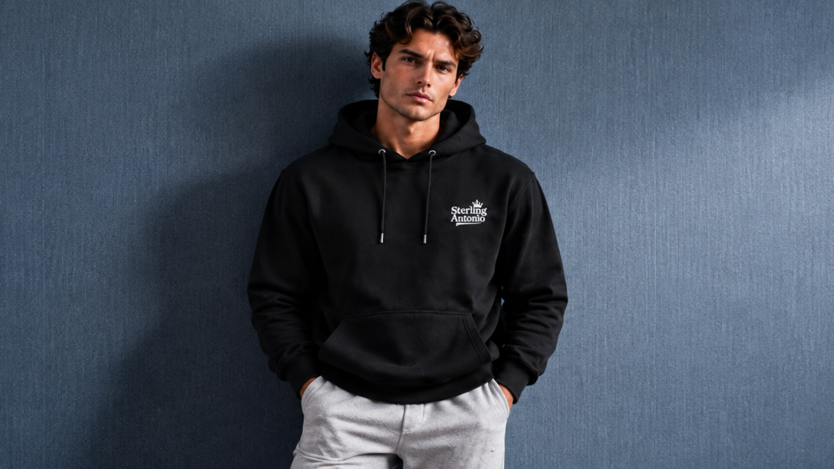 A man wearing a black pullover hoodie with a white embroidered logo on the left chest, paired with light gray sweatpants, standing against a textured blue-gray wall.