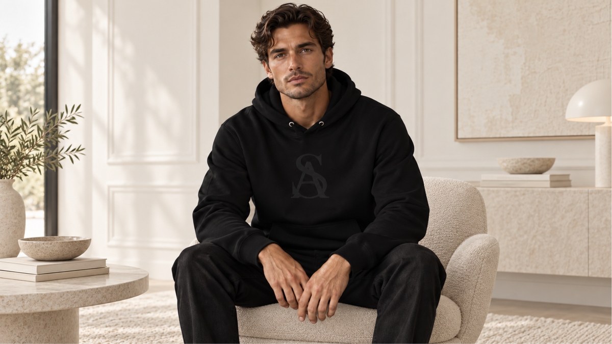 A man wearing a black pullover hoodie with a tonal dark gray logo on the chest, seated in a modern, neutral-toned living room setting.