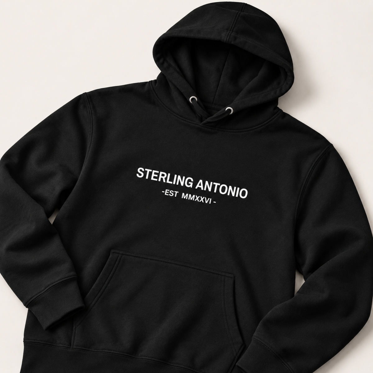 A black pullover hoodie with white text reading 'STERLING ANTONIO -EST MMXXVI-' printed on the chest, displayed on a light beige background.