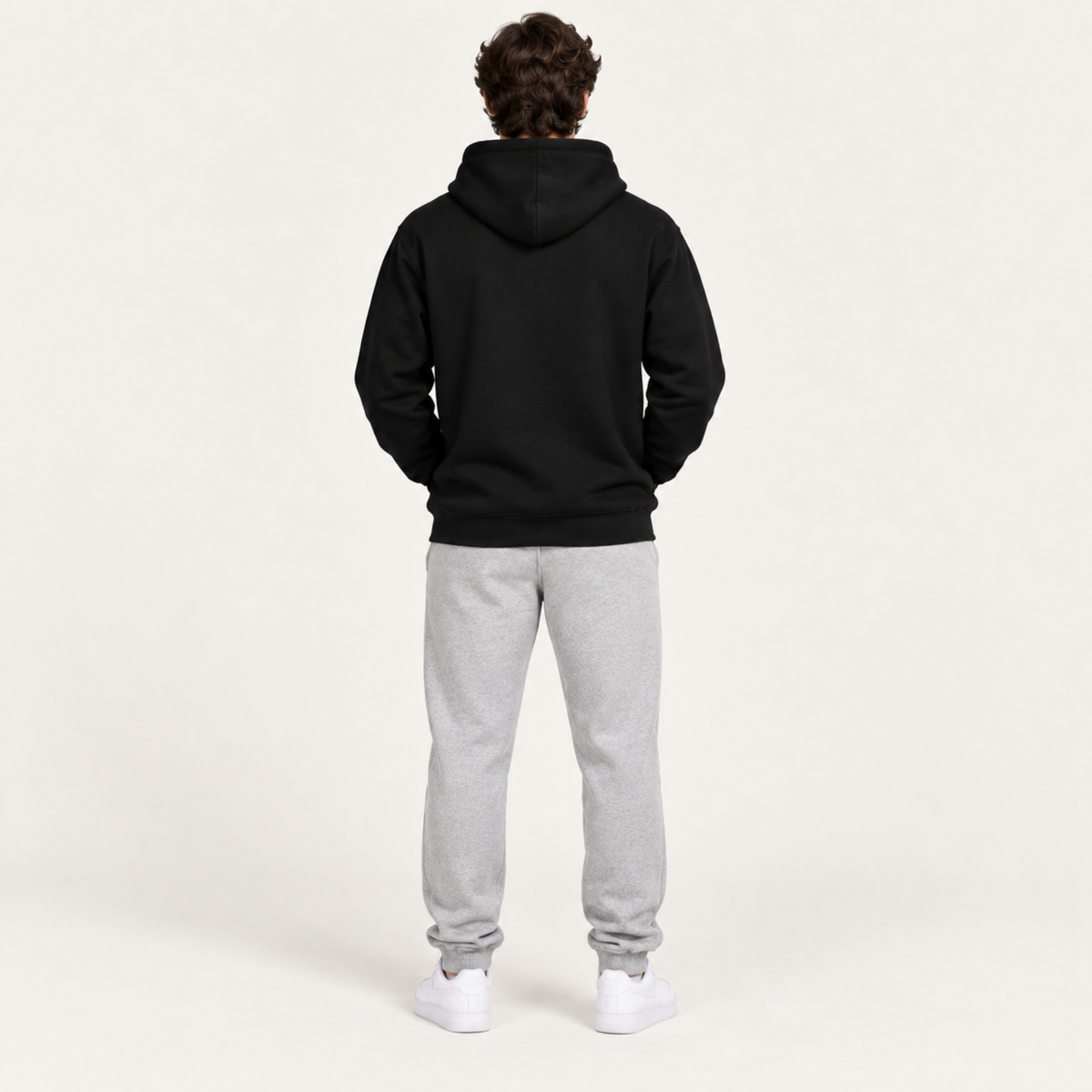 A person shown from the back wearing a black pullover hoodie, light gray sweatpants, and white sneakers against a plain off-white studio background.