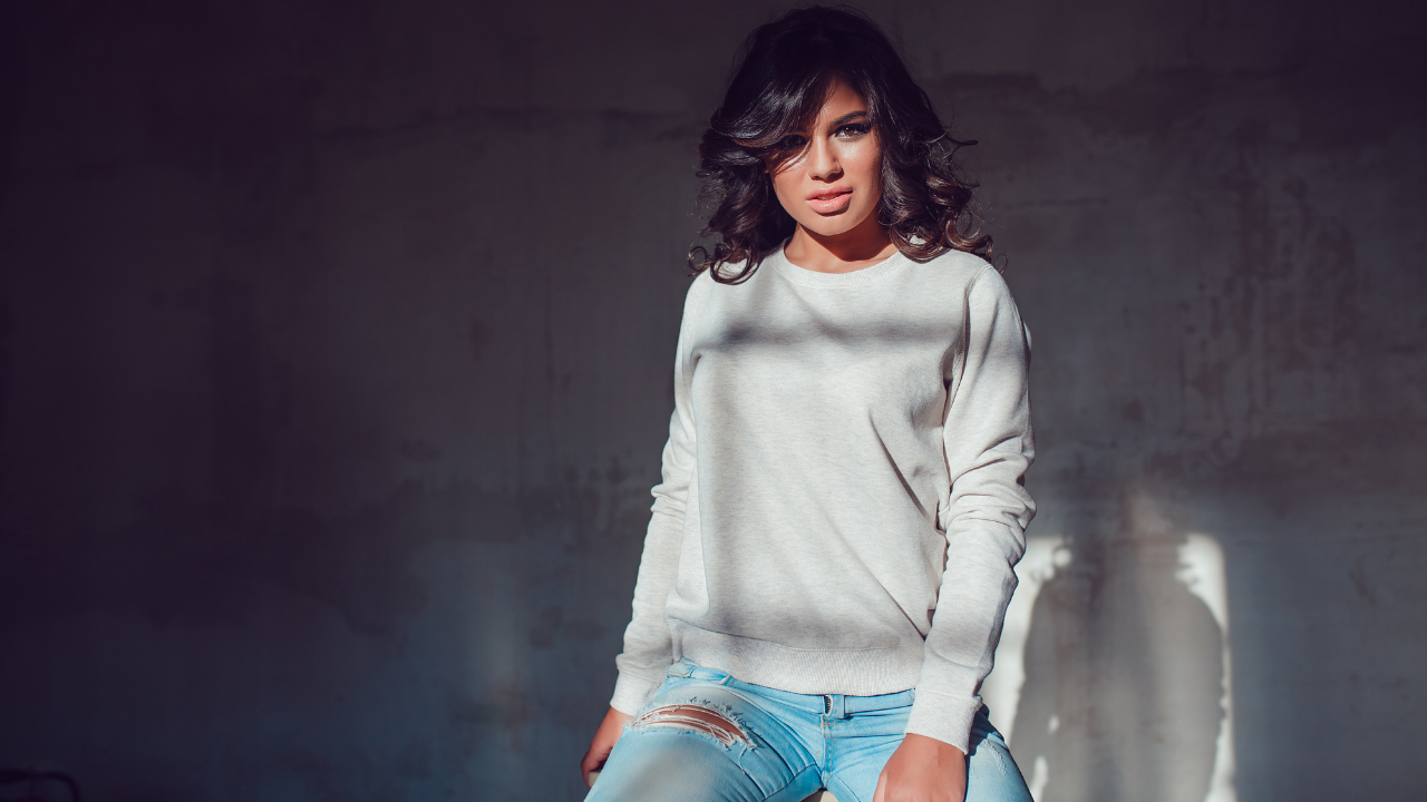 Woman wearing a white sweatshirt and blue jeans against a dark background