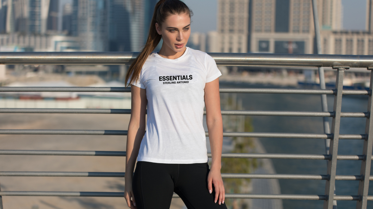 Woman wearing a white t-shirt with 'ESSENTIALS' text, standing outdoors with a cityscape background.