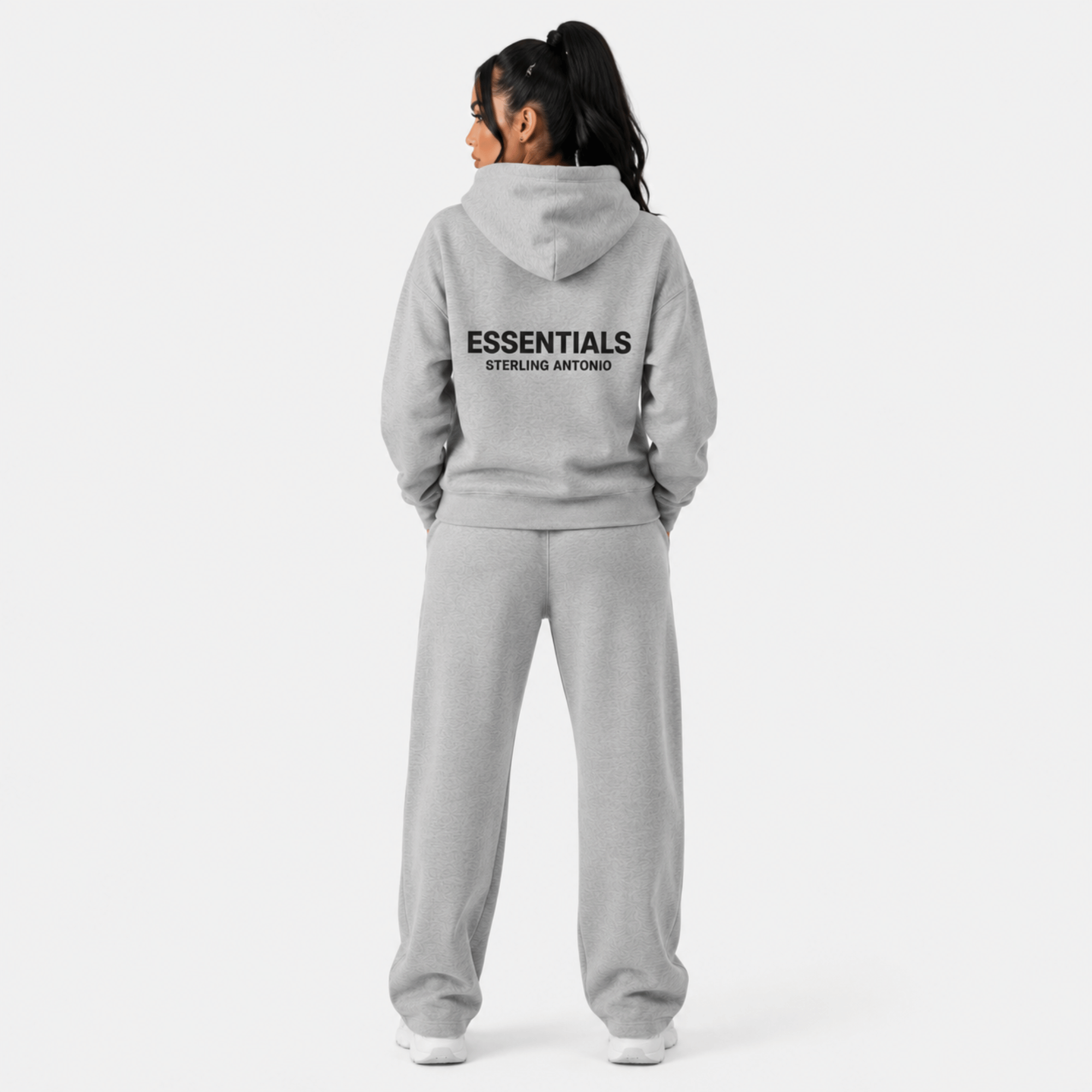 Rear view of female model wearing Sterling Antonio carbon grey essentials hoodie and matching sweatpants with oversized back logo print on light grey background