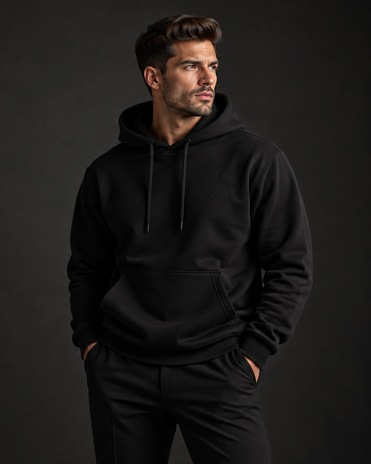 Man in The Black Label Hoodie with tonal SA monogram, three-quarter portrait on a dark background. Sterling Antonio