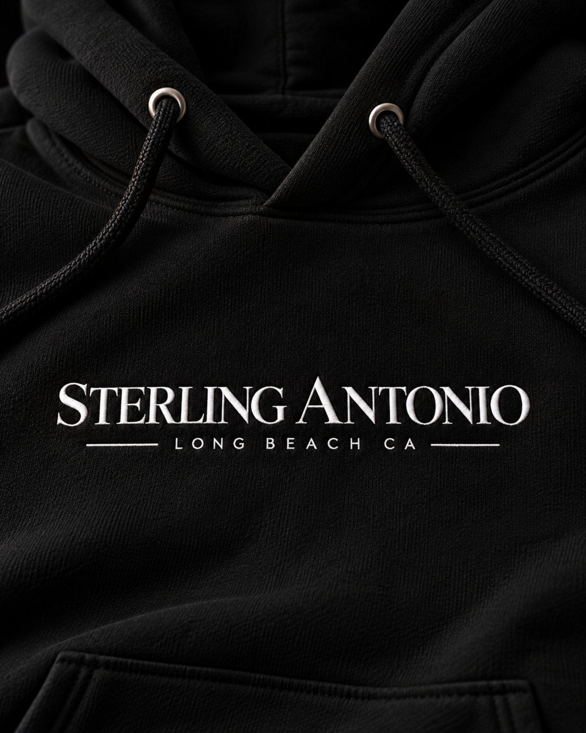 Close-up of the white Sterling Antonio Long Beach, CA wordmark on the chest of the black Founders Hoodie, with metal eyelets and braided drawcords. Sterling Antonio